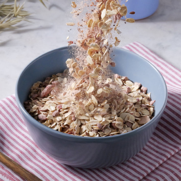 Oat cult overnight oats being poured into a blue bowl on a striped cloth