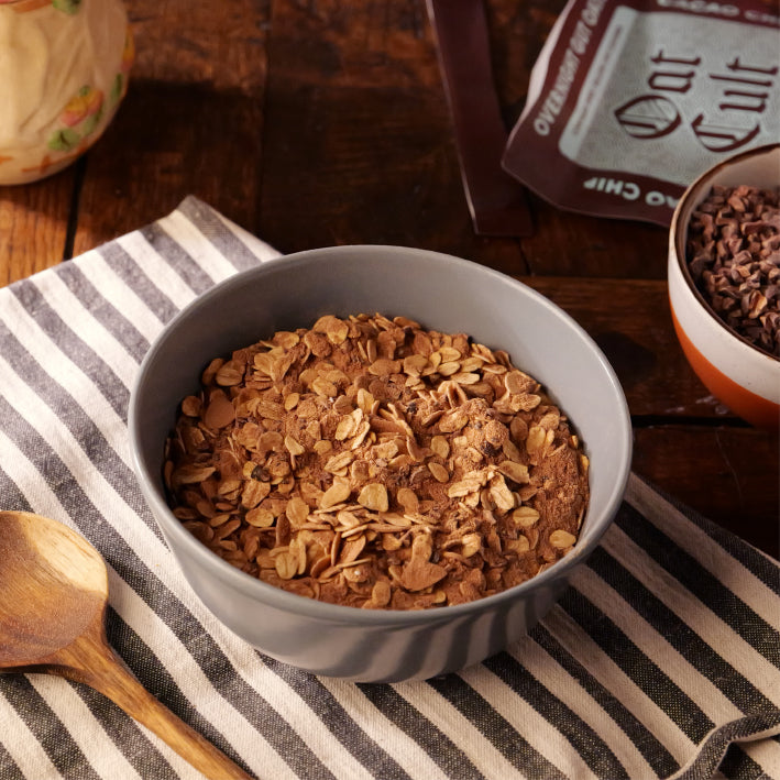 Bowl of oat cult overnight oats cacao on a striped cloth with a wooden spoon and oat milk carton in the background.